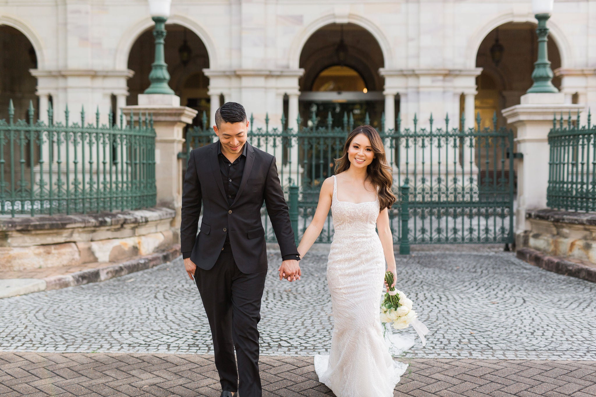 bride and groom walking through courtyard with green steel gates in front of concrete arches holding hands on their wedding day