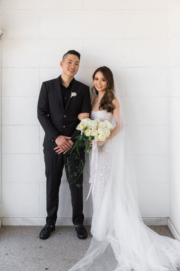 bride wearing white u-neck wedding dress holding bouquet of flowers with husband in suit standing beside her against white brick wall