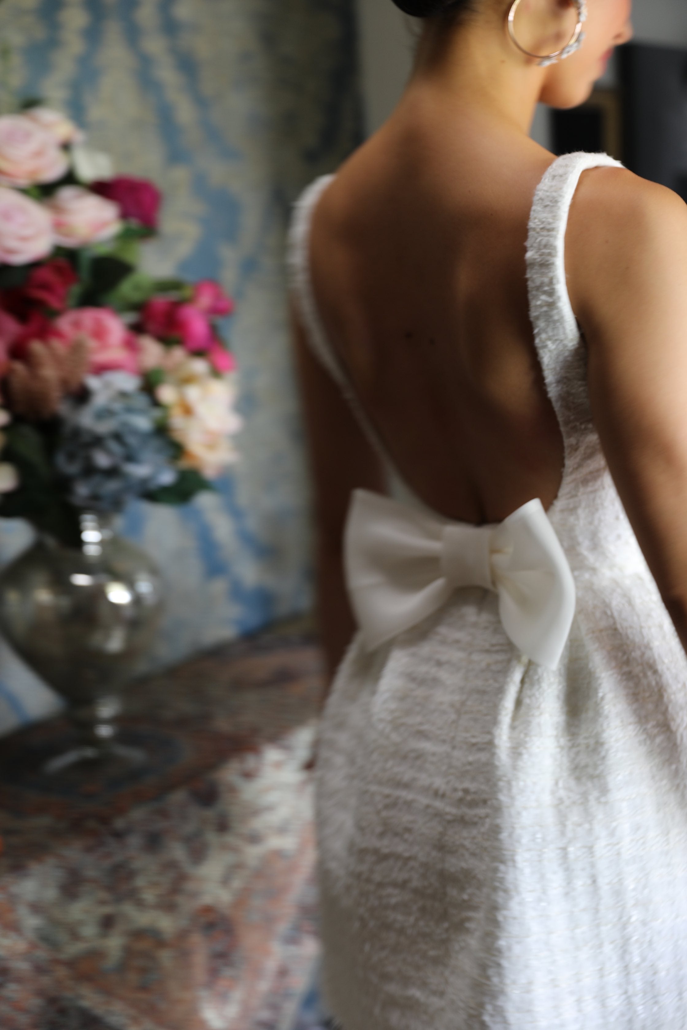 brides low back wedding dress with bow on it and flowers behind her