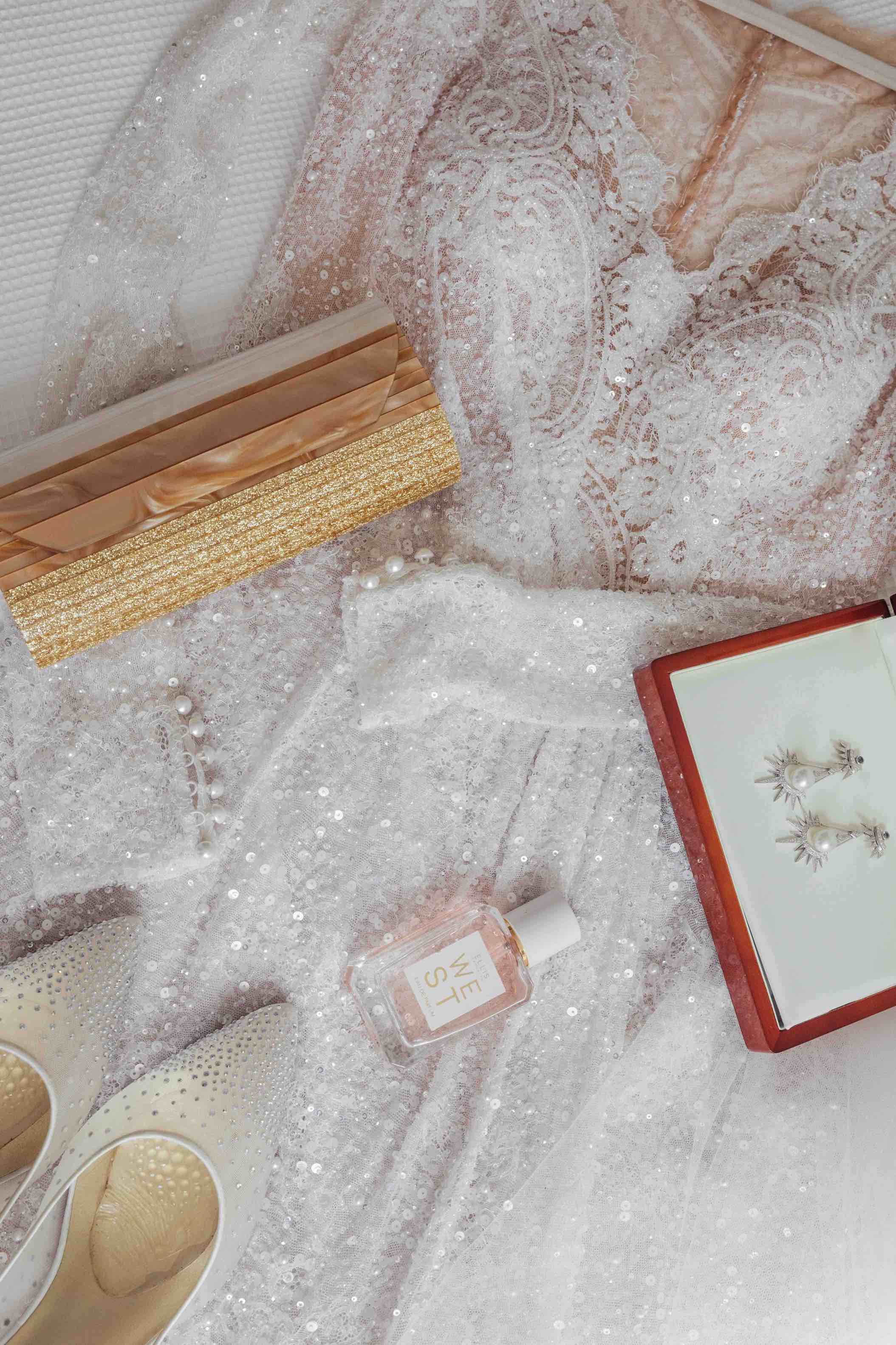 wedding dress with clutch heels earrings and perfume laid out on bed before wedding day