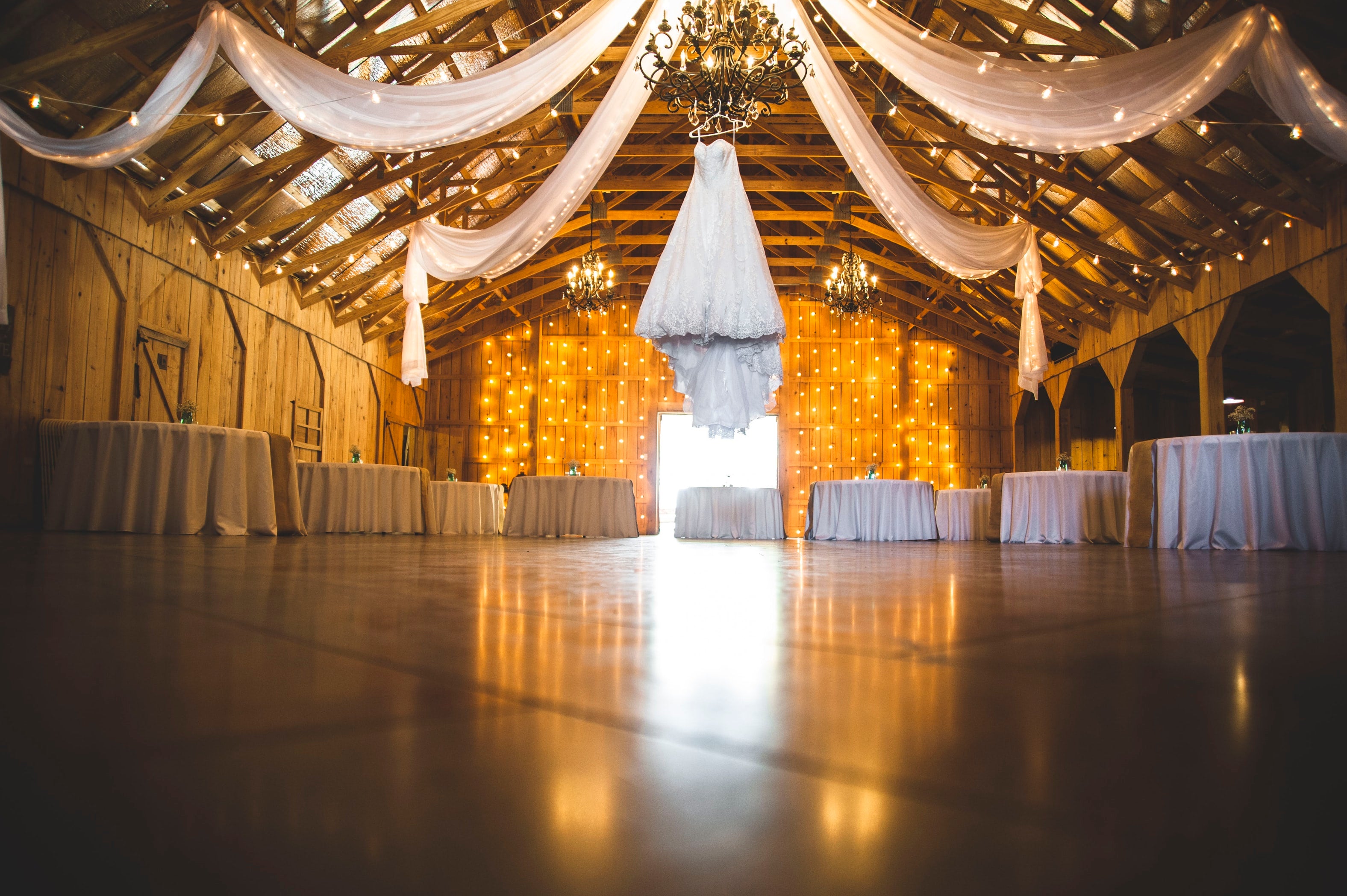 wedding venue with tables and dance floor with tulle hanging from rafters and wedding gown hanging from chandelier