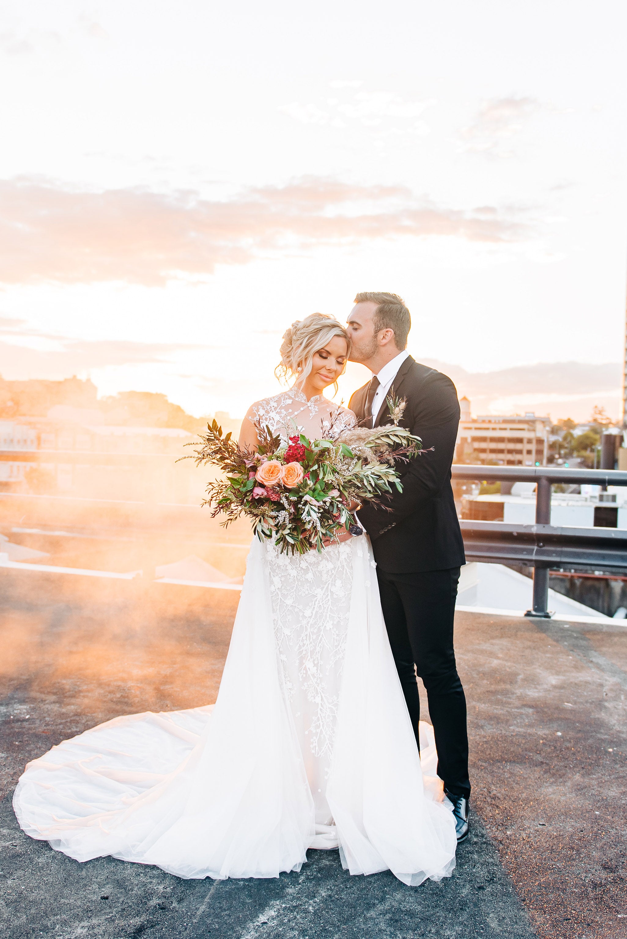 couple hugging holding flowers in wedding attire on rooftop at sunset