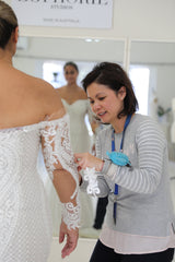 bridal designer sewing sleeve on brides wedding dress while the bride stands in front of showroom mirror
